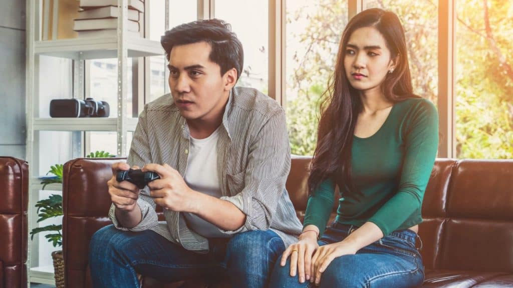 A man plays a video game next to an unhappy woman on a leather couch.