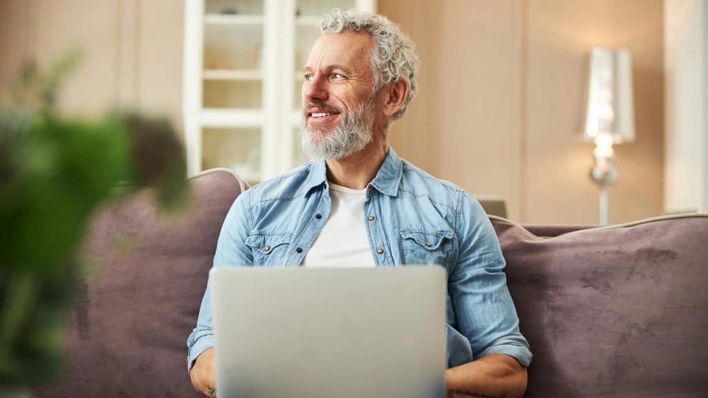 A smiling man with a beard using a laptop while sitting on a couch.