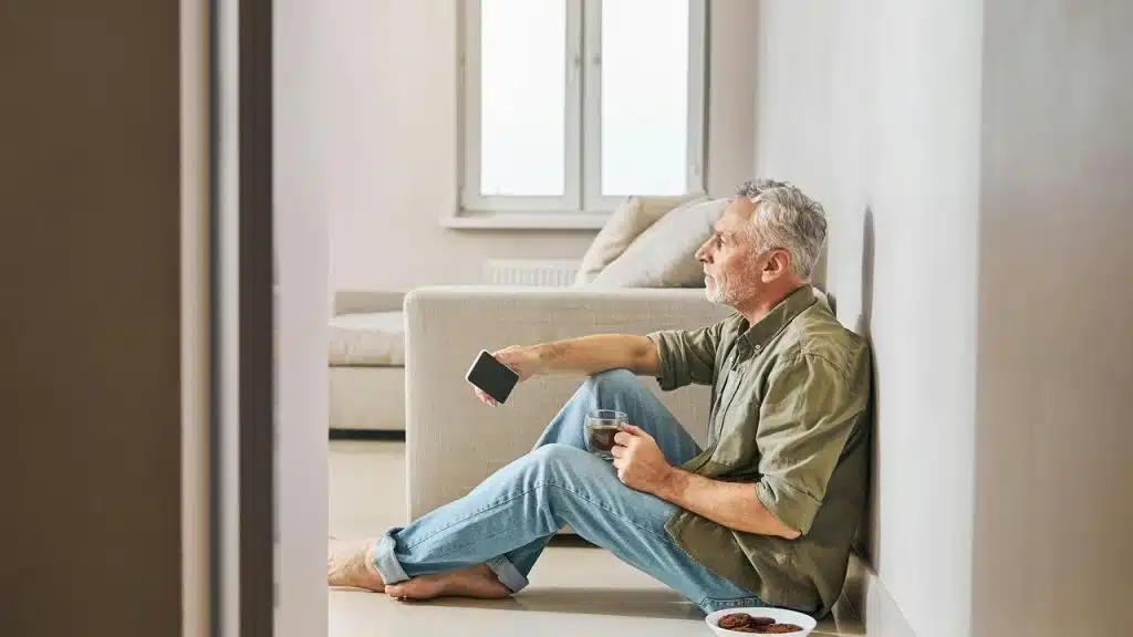 An older man sitting on the floor with a smartphone and a cup of coffee.