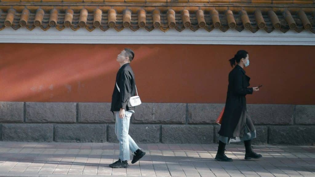 A man and woman walk in opposite directions along a street next to a red wall.