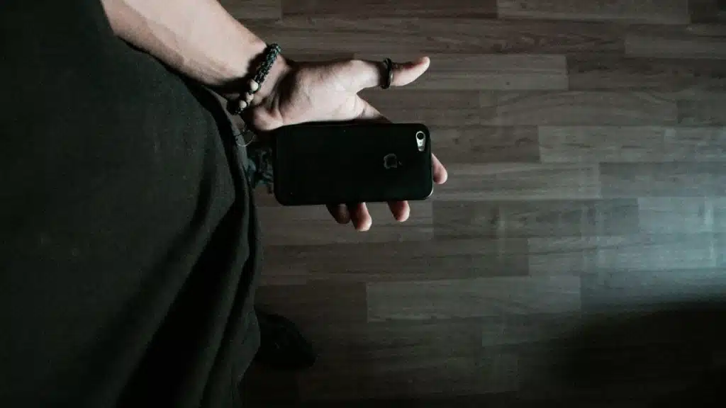 Close-up of a hand holding a black smartphone over a wood-grain floor.