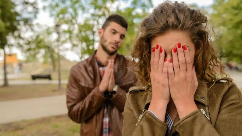 A woman covering her face while a man stands behind her with his hands clasped together.