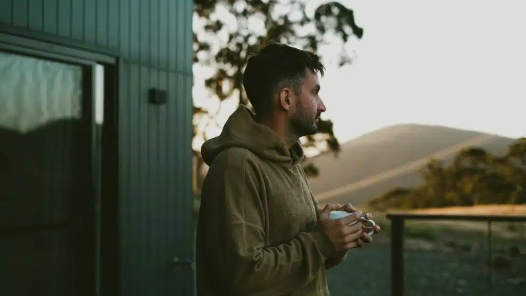 Man in a brown hooded sweatshirt holding a mug, looking out over a mountain landscape.