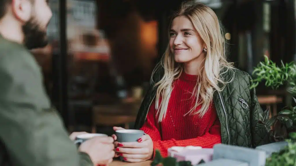 Woman smiling at a man while holding a cup of coffee at an outdoor cafe or restaurant.