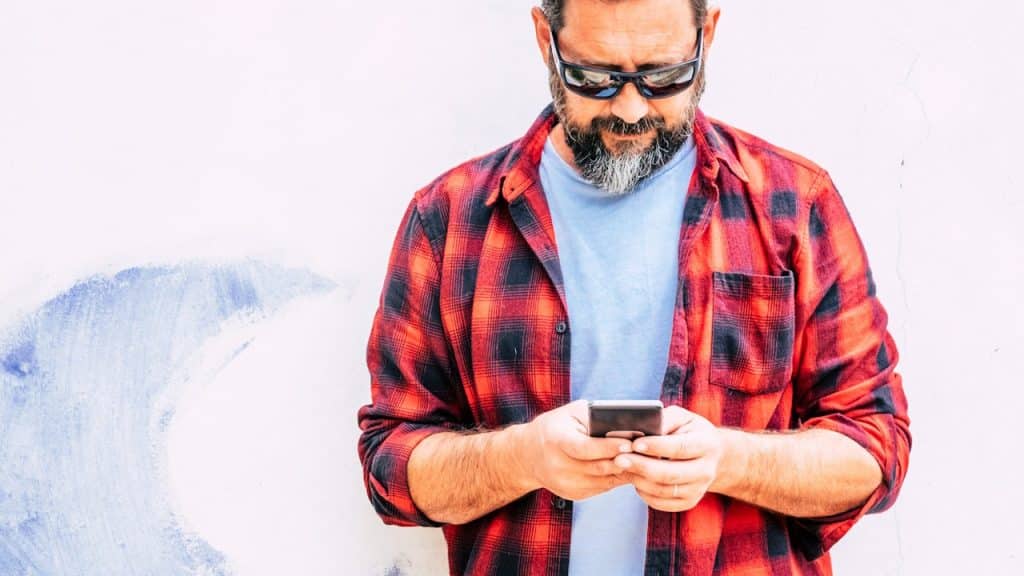 An adult man with a beard using a modern cellular smart phone standing on a white wall background.