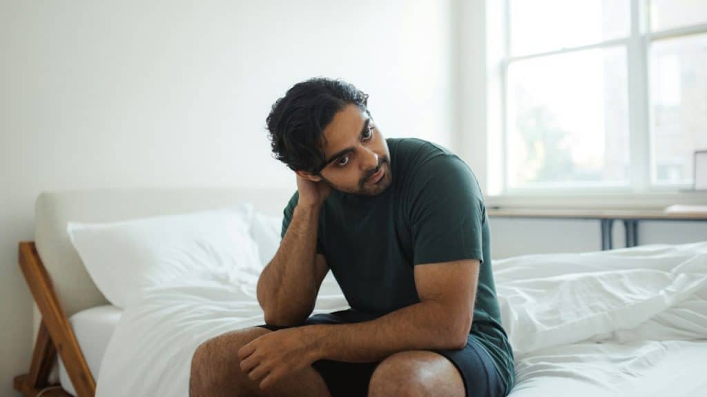 A sad man with dark hair sits on a white bed, leaning his head on his hand, looking slightly away from the camera.