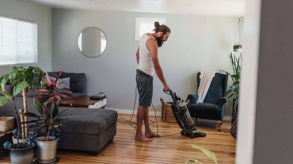 A bearded man in a tank top vacuums a wooden floor in a living room.