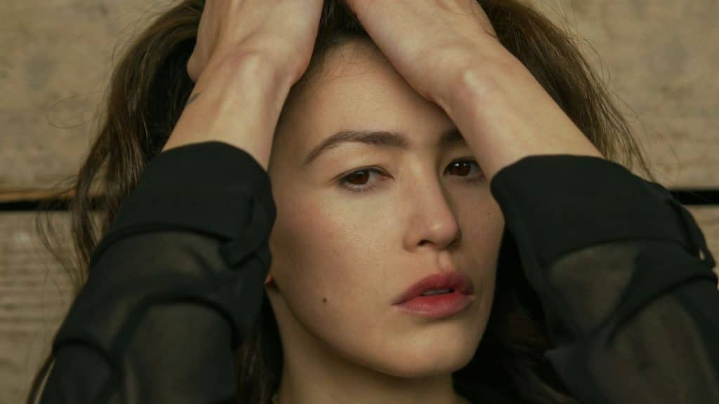 Close-up of a woman in a black sheer top holding her hands near her head.
