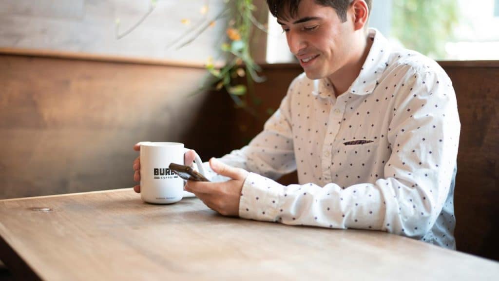 A man sitting at a café table using his smartphone and holding a coffee mug.
