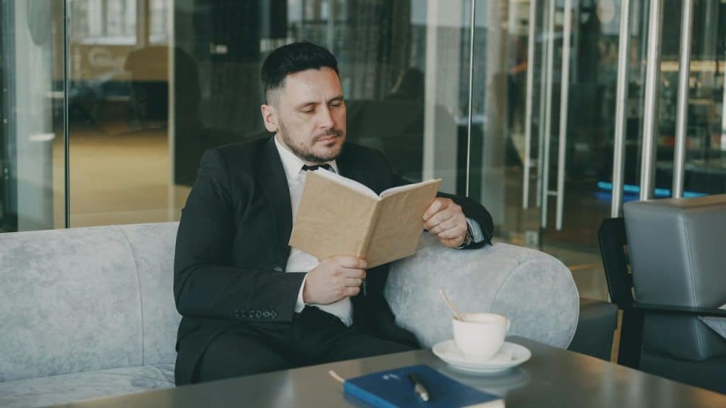Man in black suit and bow tie sitting, reading a book in a modern lounge.