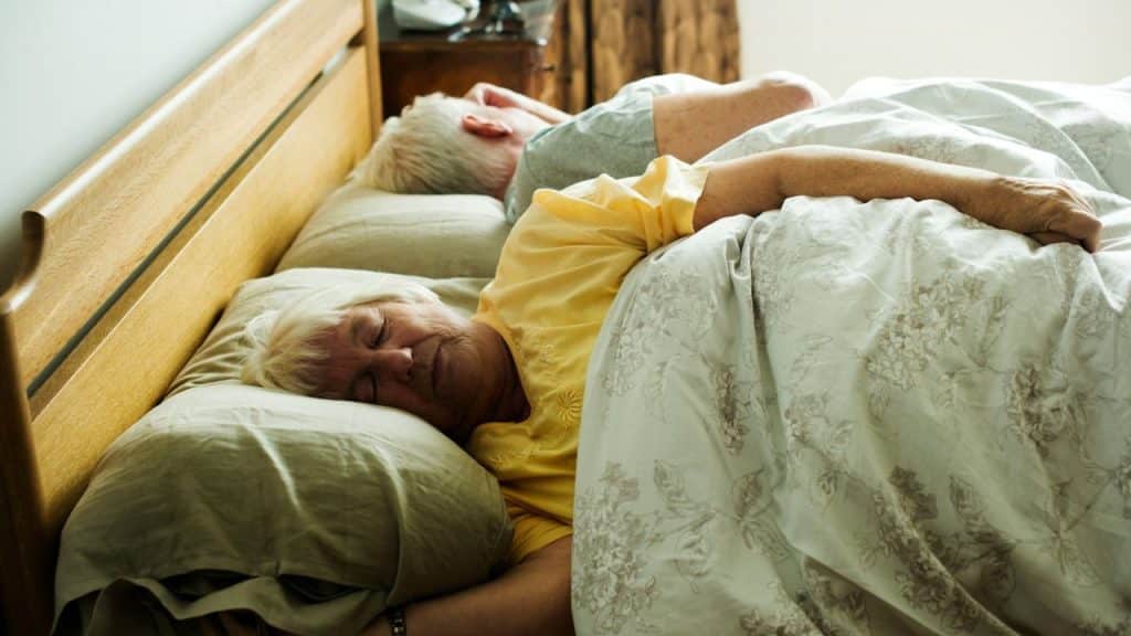 Older woman in yellow shirt sleeping on her side in a bed next to a man.