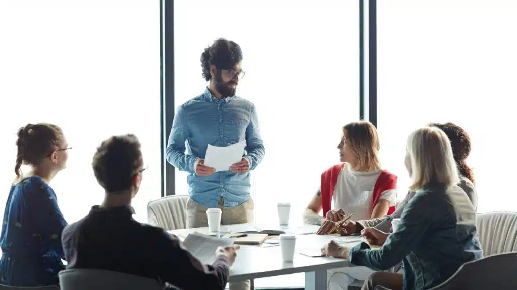 Man with beard and glasses leads a meeting, holding papers while six people listen.