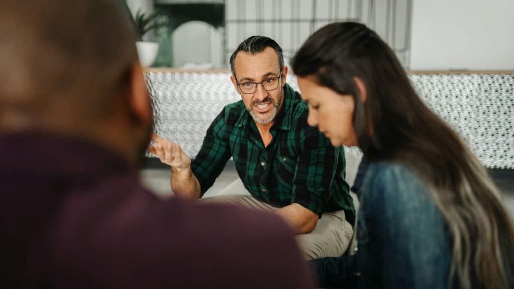 Man in glasses and a green plaid shirt talking to people in an indoor setting.
