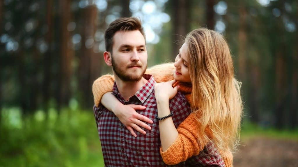 A bearded man carrying a woman on his back in a forest.