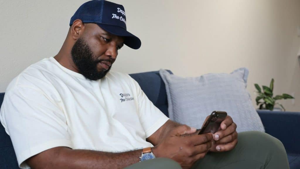 A man in a baseball cap and t-shirt sits on a couch while using a smartphone.