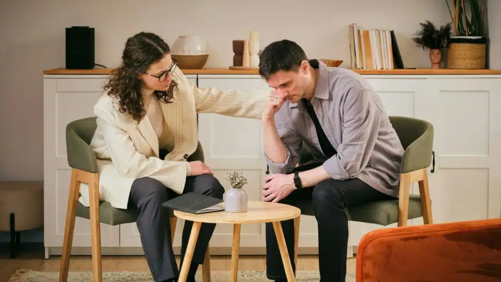A woman in a blazer rests her hand on the shoulder of a seated, downcast man.