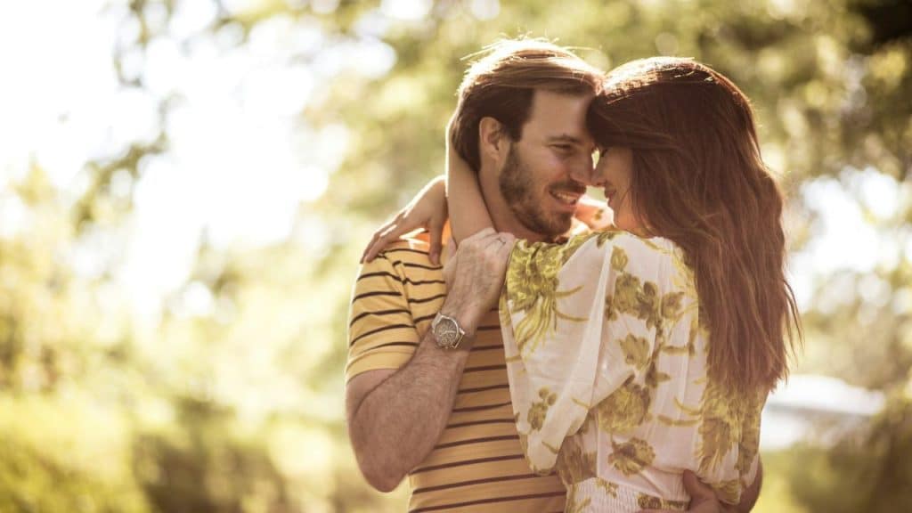 A man and a woman stand close together outdoors, embracing with their foreheads touching.