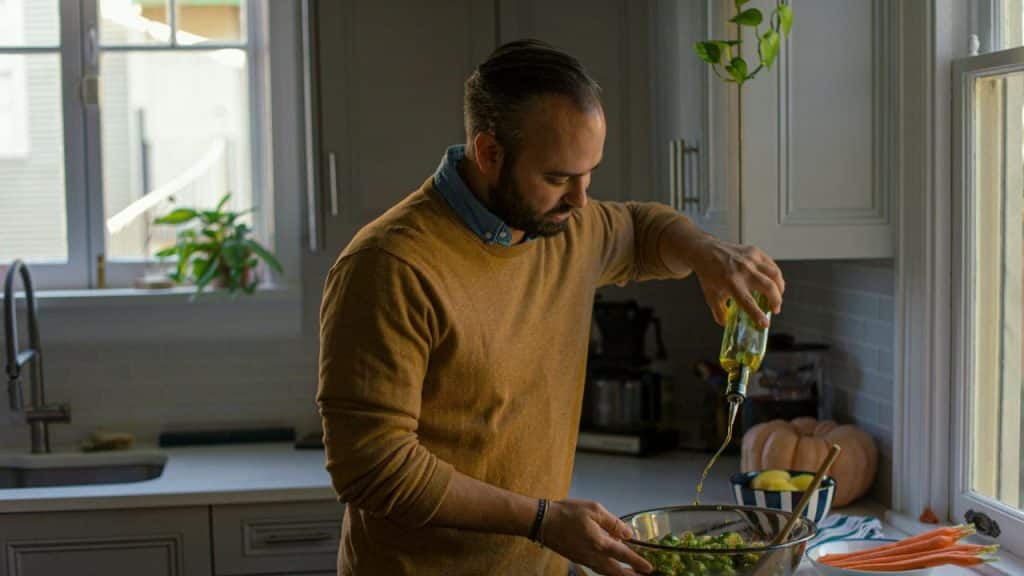 A man in a tan sweater pours oil into a bowl of vegetables in a kitchen.