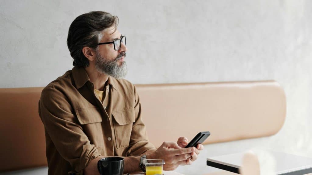 A man with glasses and a beard holds his phone while sitting at a café table.