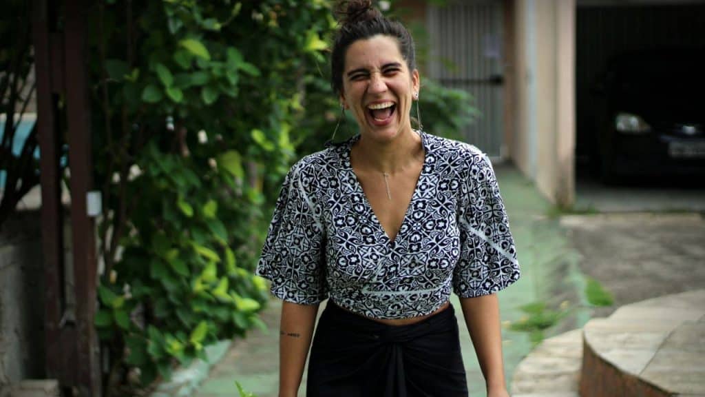 A woman in a patterned blouse laughs with her eyes closed in an outdoor setting.