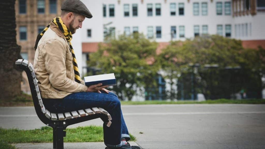 A man in a cap and scarf sits on a park bench reading a book.