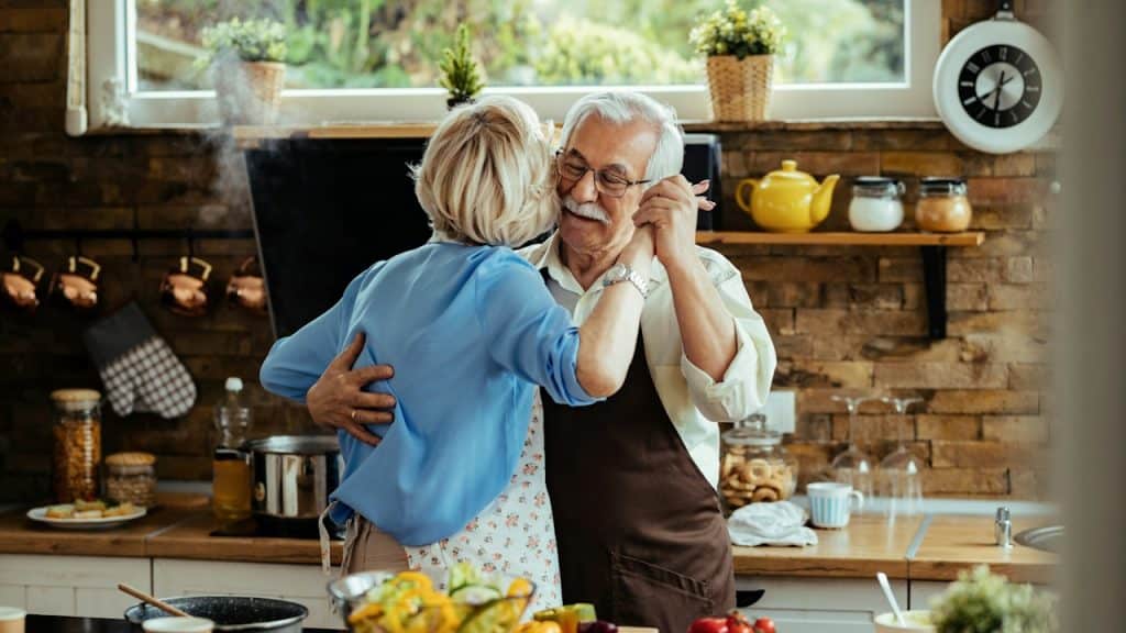 A carefree, mature couple dancing while preparing food in the kitchen.