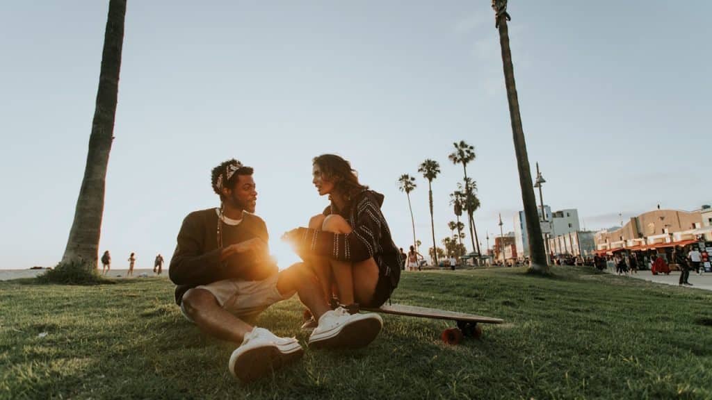 A young couple sits together on a grassy area by the beach at sunset, with the woman perched on a longboard.