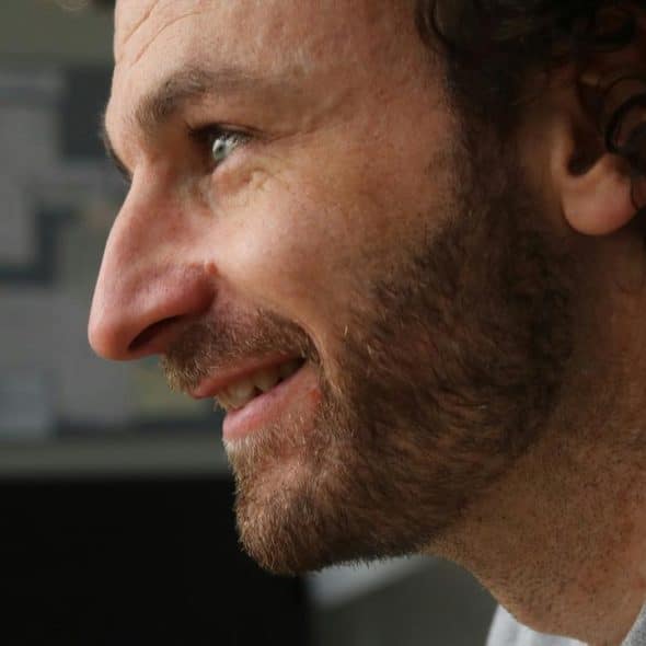 A close-up side profile of a smiling man with a beard and curly hair.