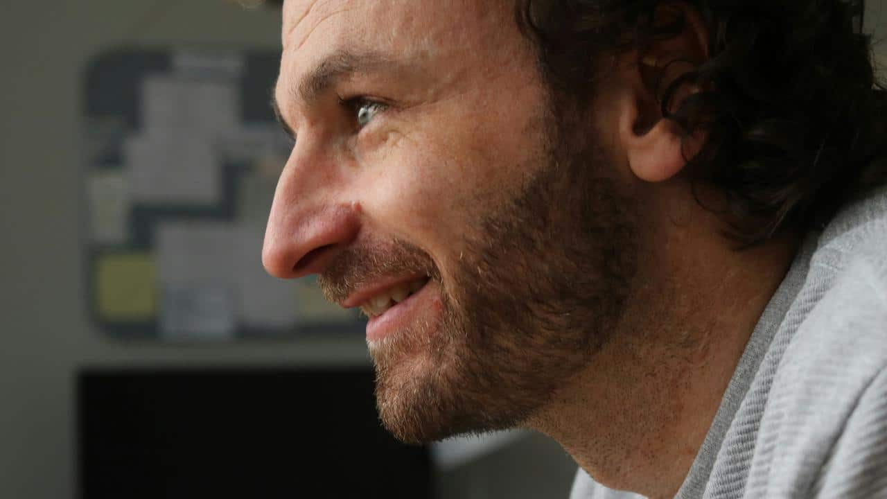 A close-up side profile of a smiling man with a beard and curly hair.