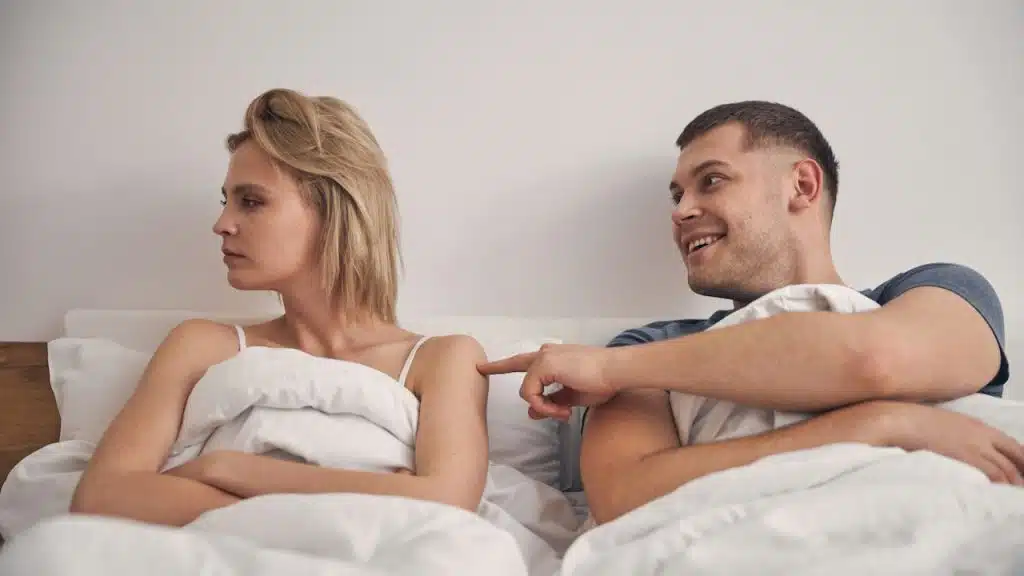 A cute blond woman looking angry in bed while a brunette male is trying to make her smile.