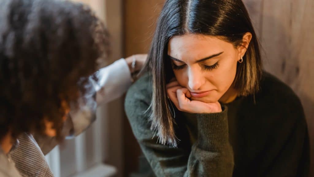 Person consoling a sad woman with short dark hair, resting her chin on her hand.