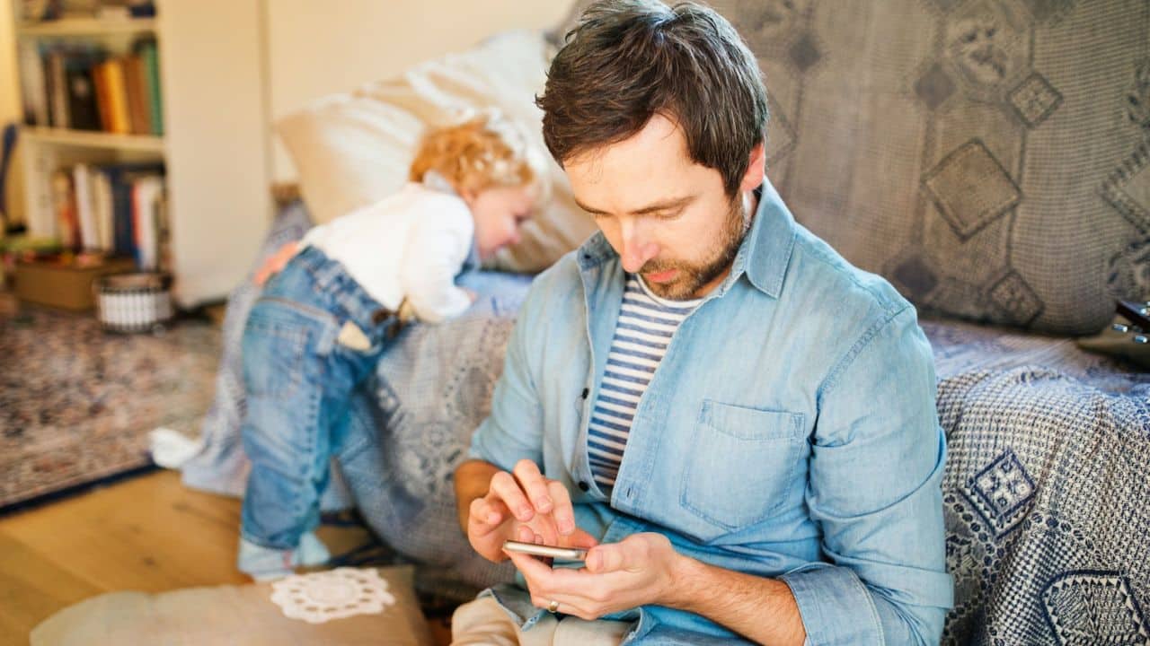 Man engrossed in his phone while a small child plays in the background behind him.