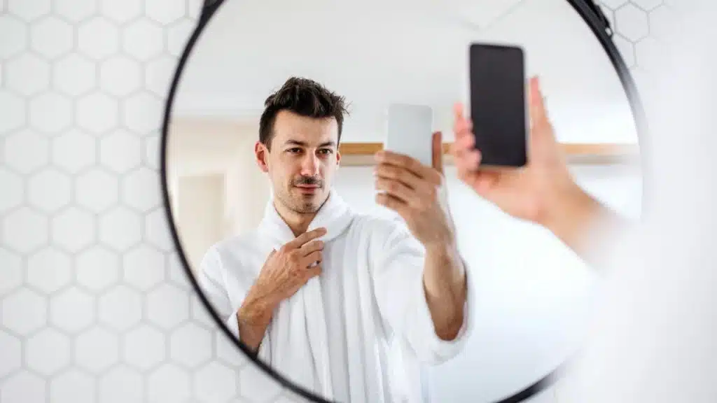 Man in a white bathrobe taking a mirror selfie with his smartphone in a bathroom.