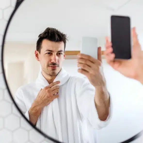 Man in a white bathrobe taking a mirror selfie with his smartphone in a bathroom.