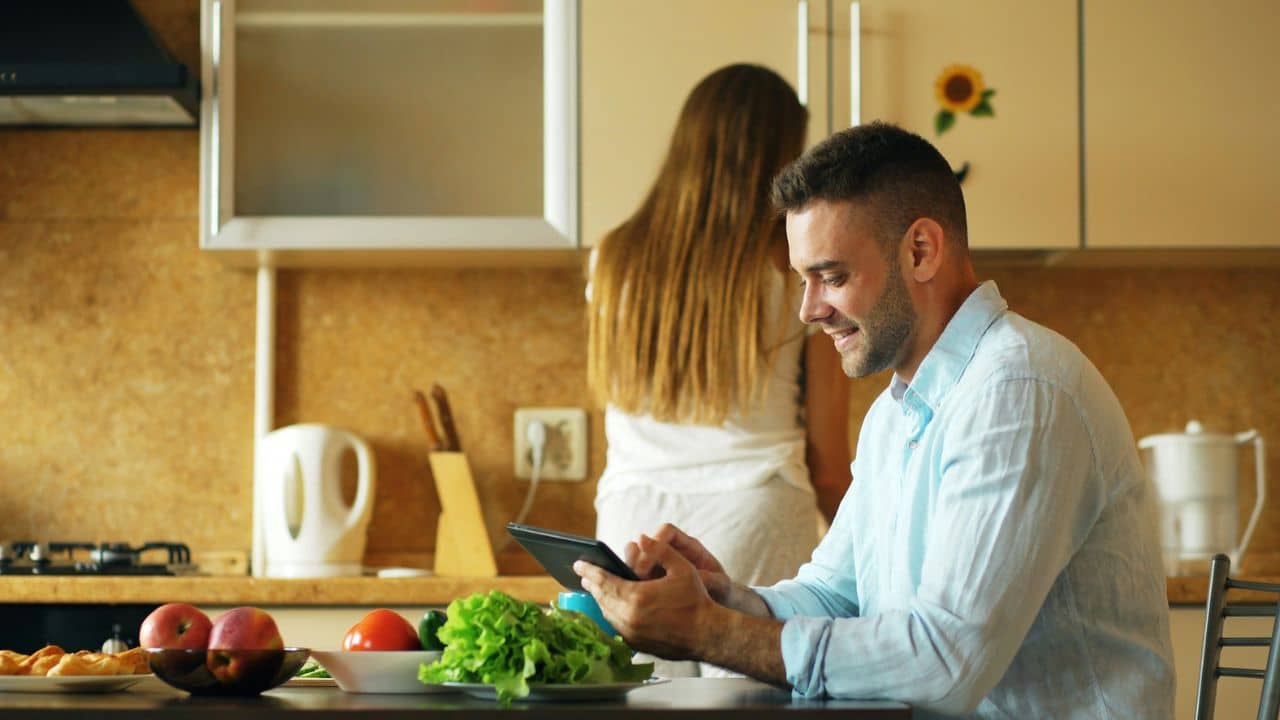 A man smiles while using a tablet at a kitchen table; a woman stands behind him.