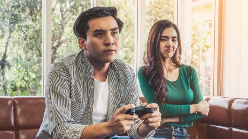 A man plays a video game on a couch while a woman next to him gives a disapproving look.