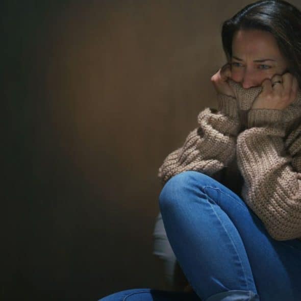 A distressed woman in a bulky sweater and jeans sits, holding her hands to her mouth.
