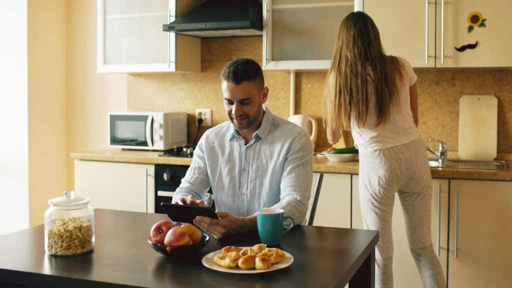 A man uses a tablet at a table while a woman attends to the kitchen counter.