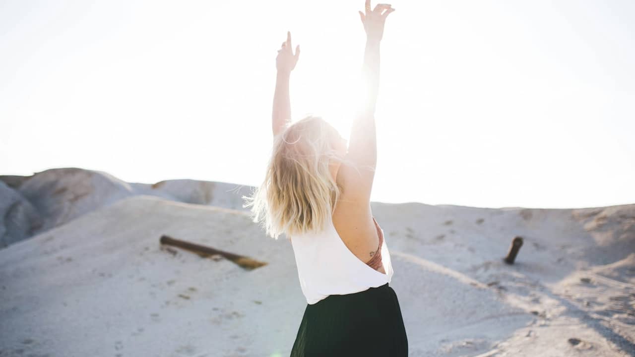 A woman with blonde hair stands in a sandy, desert-like landscape with her back to the camera and her arms raised toward the bright sky.