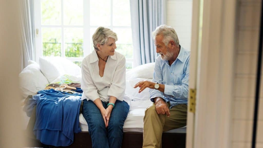 Elderly couple sitting on a bed in a sunny room, having a conversation.