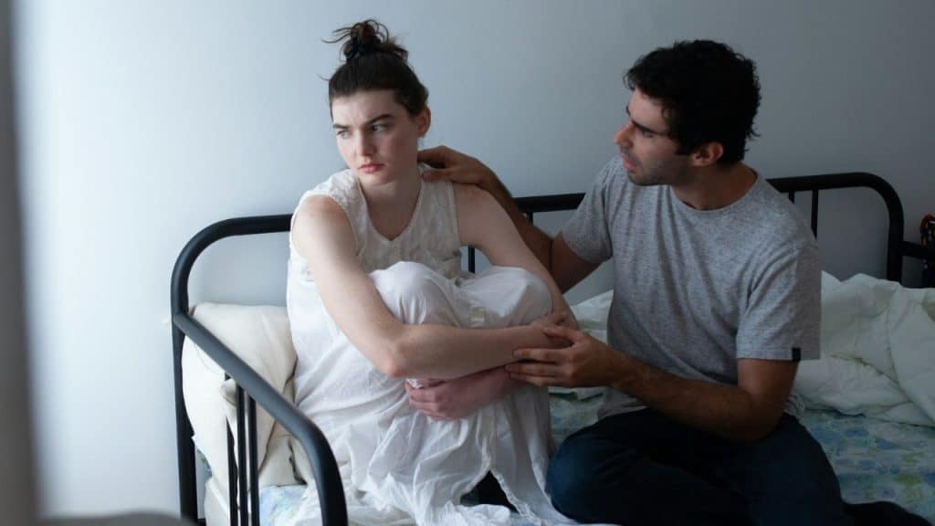 A man in a gray shirt comforts a woman in a white nightgown on a metal bed.