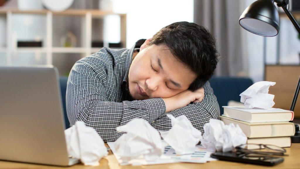 Man asleep at a desk surrounded by crumpled paper, books, and a laptop.