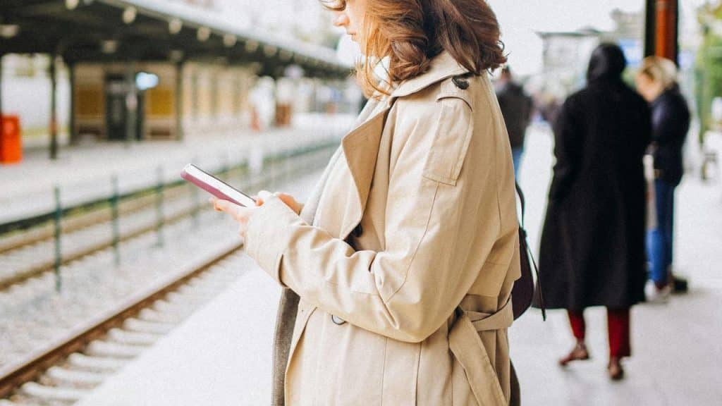 A person in a beige coat using a phone while waiting at a train station