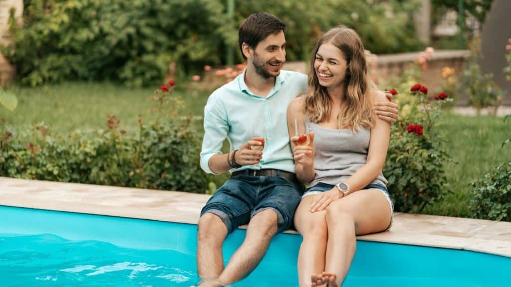A man and a woman sitting on the edge of a pool, smiling in a garden setting.