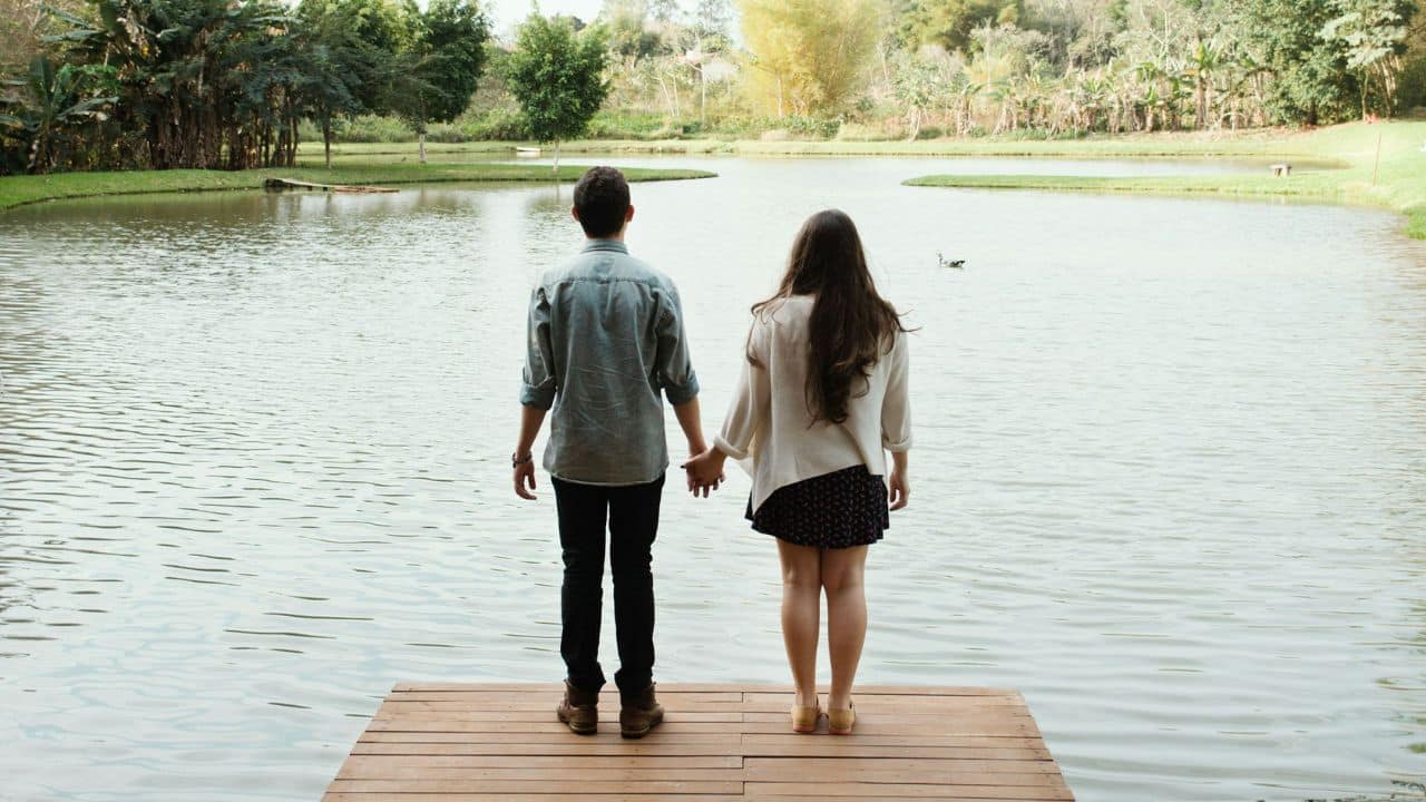 A couple standing on a wooden dock, holding hands and looking out over a lake.