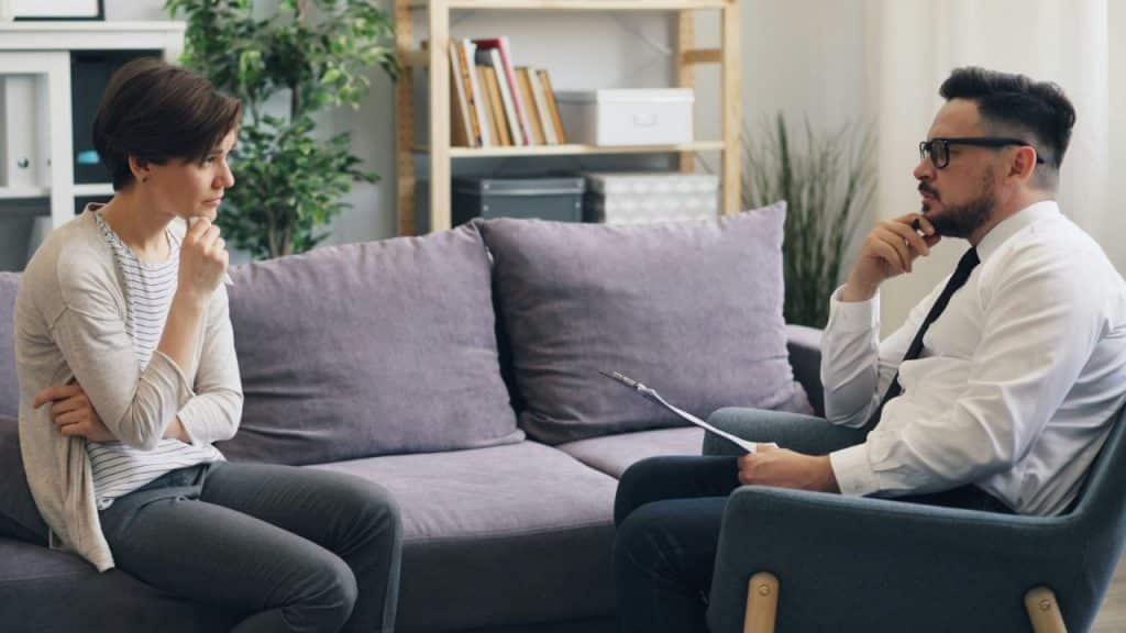 Woman sitting on a couch talking with a man holding a folder in a couch.