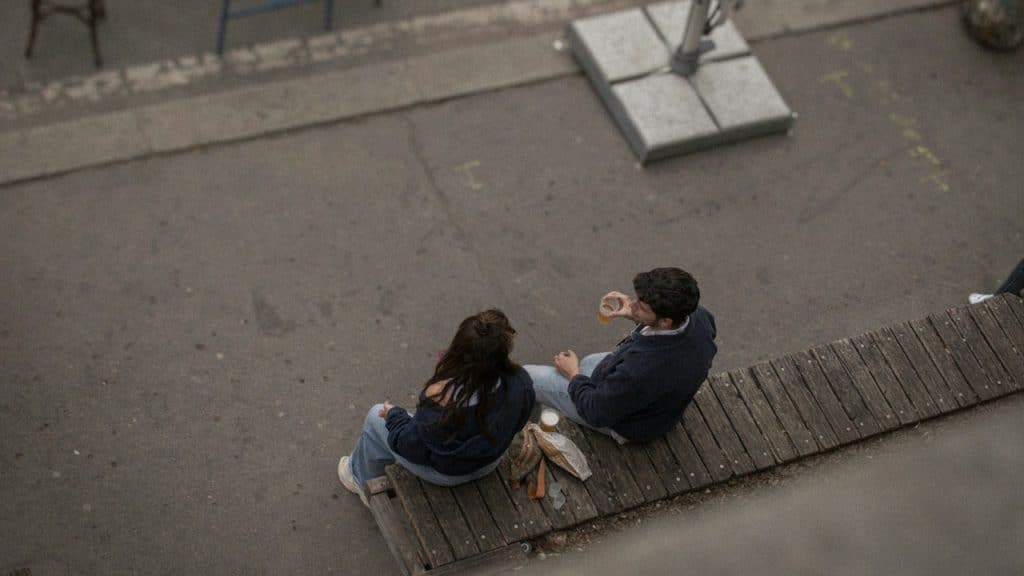 Two people sitting on a wooden platform near a street, viewed from above.