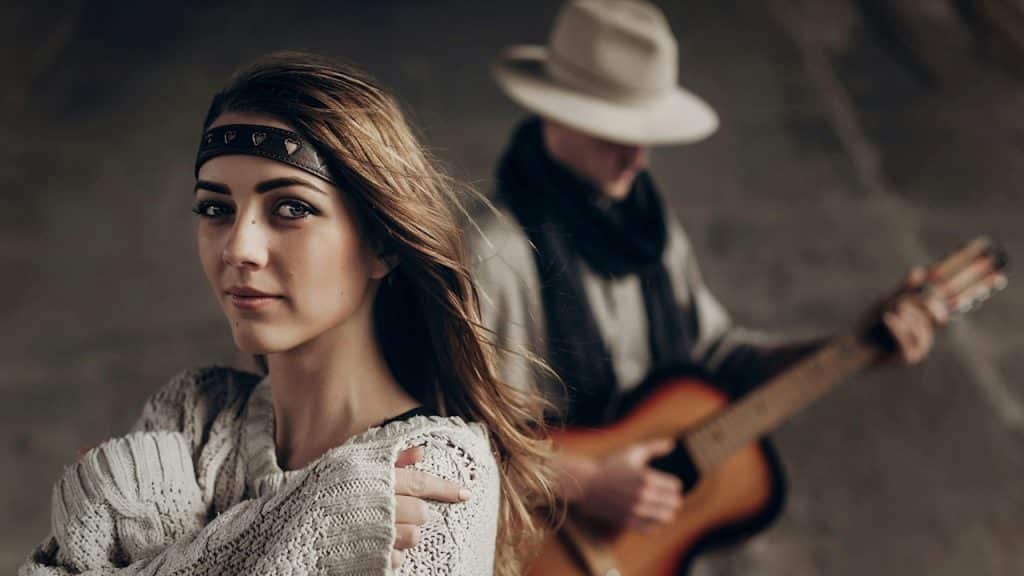 A beautiful, sensual, indie girl in hipster-boho clothes posing in front of a handsome cowboy guitar player.