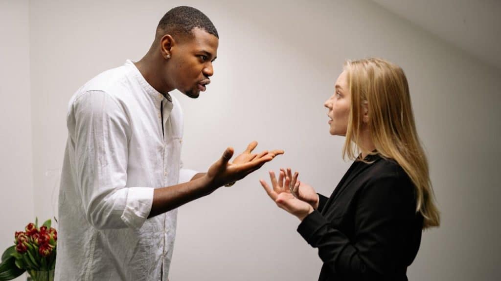 A man and woman are arguing; the man gestures with open hands.