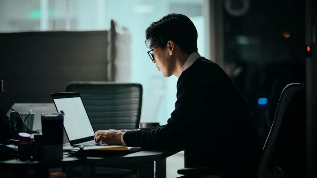 Asian man in glasses and dark sweater works on a laptop in a dimly lit office.